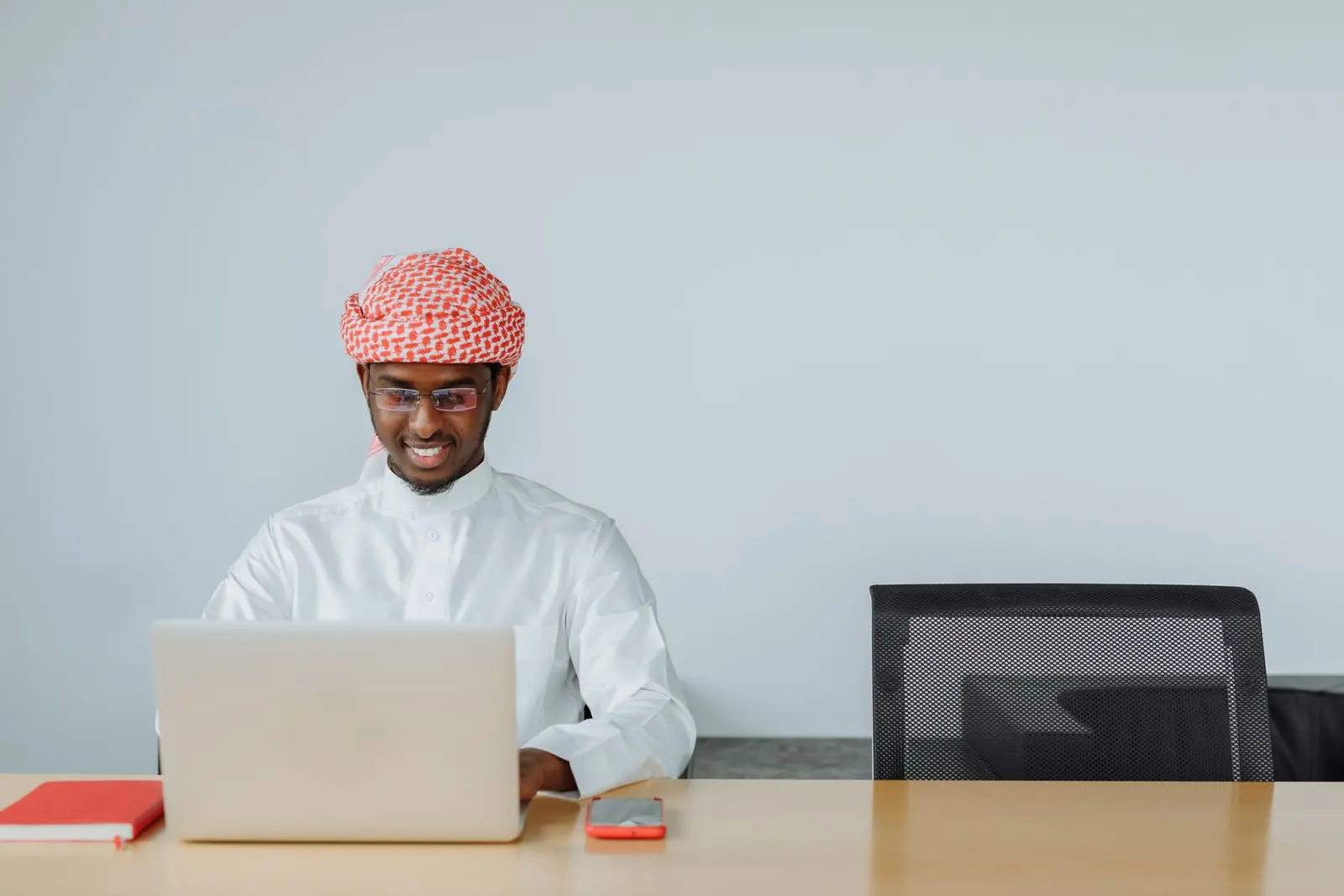 Smiling young man in a white thobe and shemagh studying on his laptop